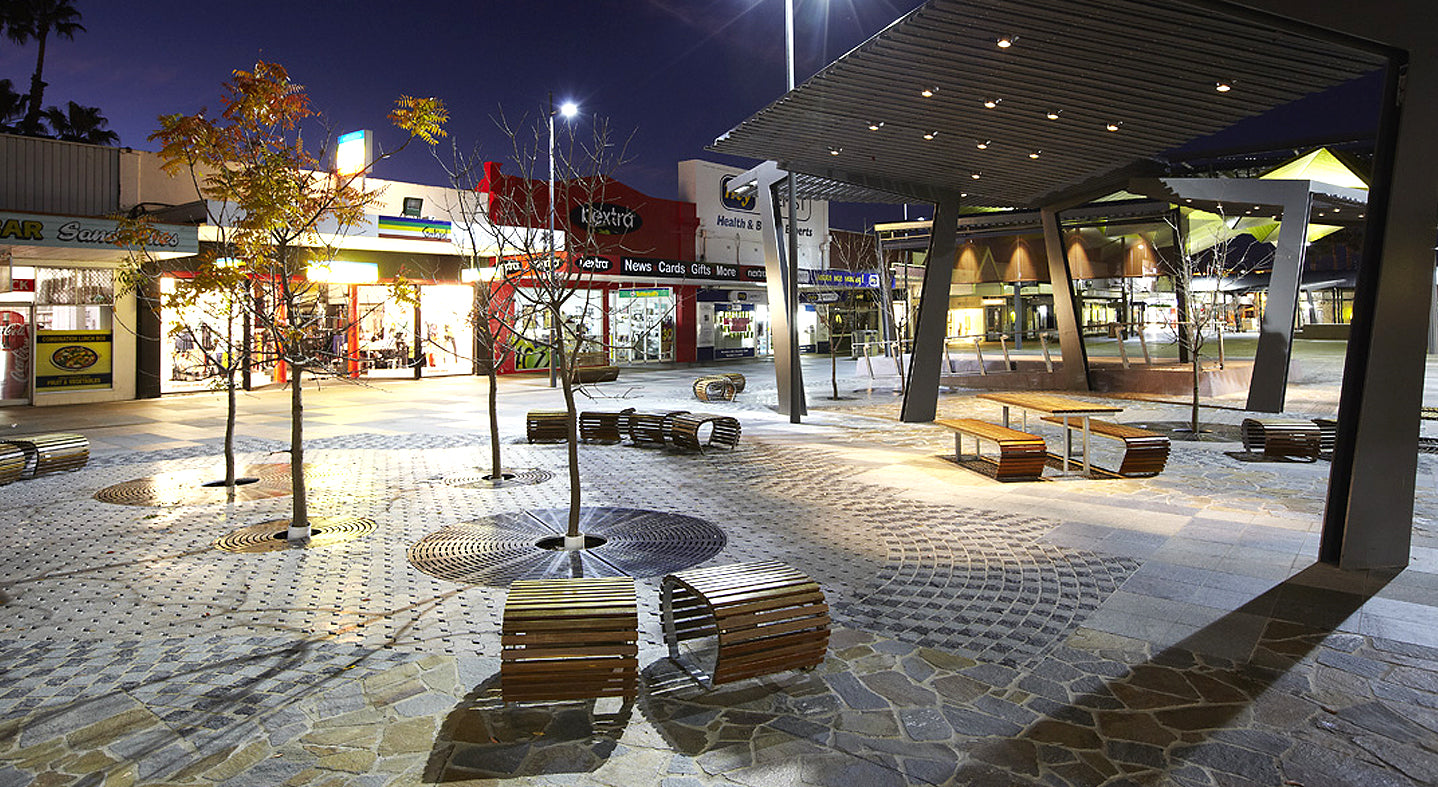 Evening view of a shopping center with illuminated storefronts and outdoor seating area.