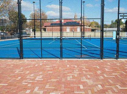 Blue tennis court behind a black chain-link fence with a brick pathway in the foreground.